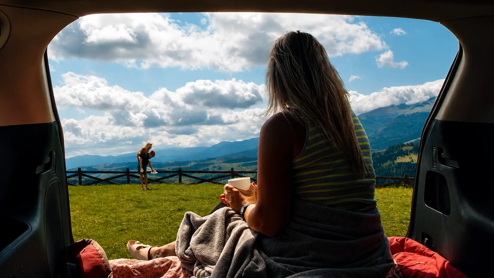 Woman wrapped in a blanket in an open car boot, looking out over a mountain valley with a mug of coffee