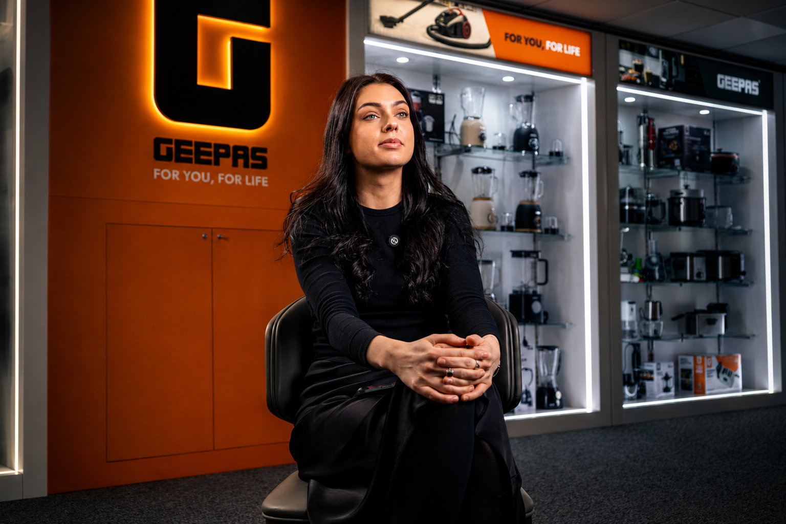 Woman seated in front of an illuminated Geepas sign with product display cabinets behind her