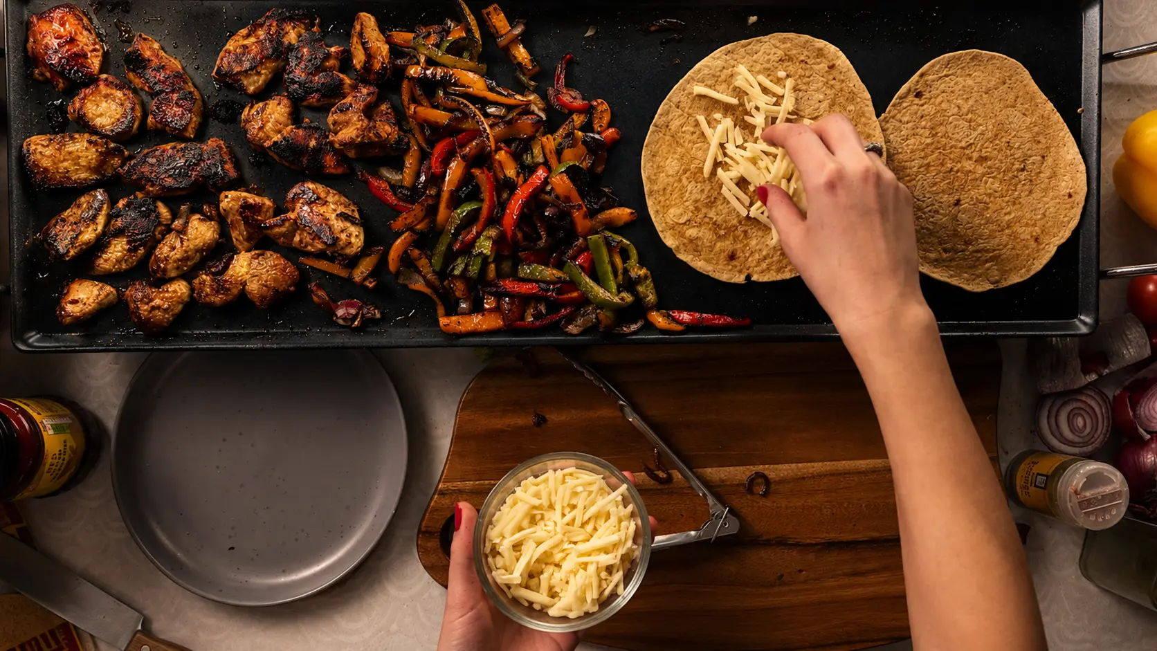 Top-down griddle shot of charred chicken, peppers and tortillas being topped with cheese